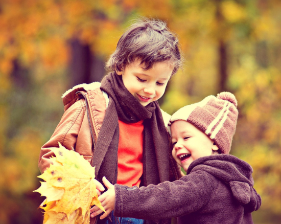 Two laughing children in warm autumn clothing holding up a large yellow maple leaf, with colourful autumn trees in the background